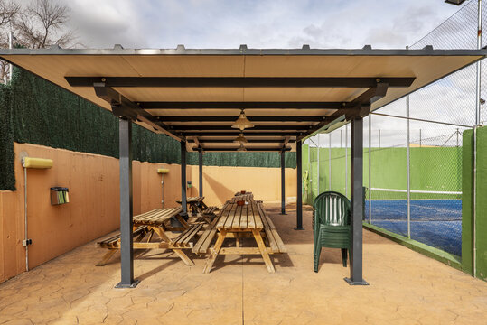 A metal and wood frame gazebo with long wooden tables facing a tennis court with a blue indoor painted floor