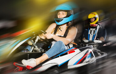 Fototapeta na wymiar sportive adulr woman in helmet driving car for karting with other people in sport club indoor