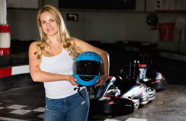 Fototapeta na wymiar Smiling glad positive woman with helmet standing near cars for motor racing in sport club