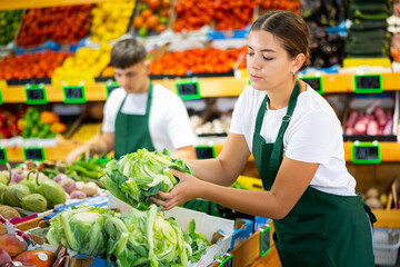 Supermarket female employee lays out fresh cauliflower on shelves in greengrocery store