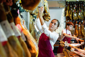 Skilled interested young girl, owner of butcher shop selling delectable dried Iberian jamon working...