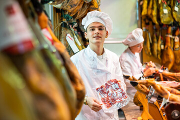 Confident positive young salesman in white uniform standing near counter in butcher shop, offering package of sliced delicious jerky Iberian jamon