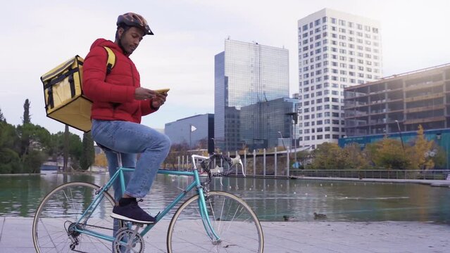Portrait of a happy delivery man who is sitting on a bicycle and texting on the smartphone. He is carrying a yellow backpack for delivery on his shoulders. High quality 4k footage