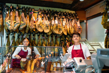 Fototapeta na wymiar Positive girl and guy, sellers of small local butcher shop specializing in sale of jerky Iberian jamon working together behind counter