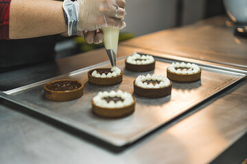 A baker wearing transparent glove decorating tarts placed on a baking tray with white cream using a pastry bag. Blurred background. High quality photo