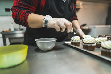 A baker wearing red plaid shirt, black apron ad transparent gloves decorating cupcakes with white cream placed on a baking tray in the kitchen. High quality photo