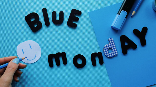 Chica con u&ntilde;as azules dibujando una cara sonriente en un fondo azul con letras de Blue Monday y material de papeler&iacute;a azul. Lunes azul alegre	