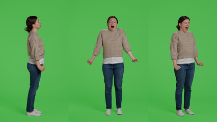 Fototapeta premium Tired female model yawning on studio camera, posing over green screen backdrop. Young adult being exhausted and sleepy standing over isolated full body greenscreen background, falling asleep.