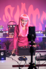 Dj with pink hair recording music session with smartphone camera while playing song at mixer console, standing in audio studio over pink background. Artist checking sound , celebrating night life