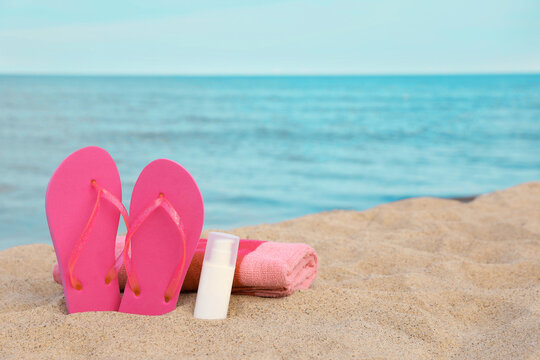 Beach towel, slippers and sunscreen on sand near sea, space for text