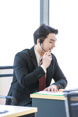 Closeup shot of Millennial Asian young professional successful male businessman operator in formal suit with stereo headset microphone sitting working talking with customer in office