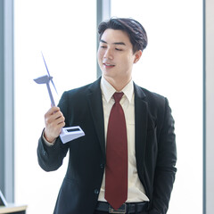 Millennial Asian young professional successful male businessman manager in formal suit standing smiling holding windmill model showing thumb up in company office while colleague working at workplace