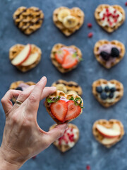 Hand holding a strawberry topped heart waffle, and others in soft focus below.