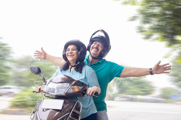 Happy senior indian couple wearing helmet riding motor scooter on road. Retirement life, Adventure...