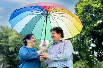 Happy senior indian couple standing under big multicolor or colorful umbrella at summer park....