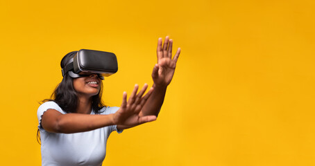 Young indian woman wearing virtual reality simulator gesturing on yellow background. Horizontal banner.