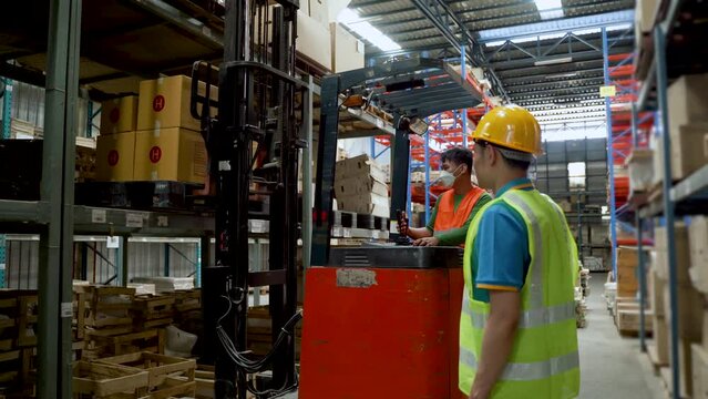 4K, Forklift driver controls forklift to load pallets on racks in a large warehouse. Colleagues advised to be careful of nearby accidents and to wear safety vests. Check products stored in shelves.