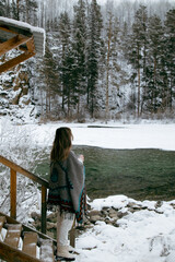 Young woman standing outdoors in winter by a mountain stream. She is dressed warmly and looks serene and contemplative. Snow-covered surroundings and flowing water create a calm, natural, and pictures