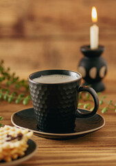 A black cup of coffee on a wooden brown background, a plant and a waffle dessert next to it