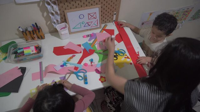 An Asian female teacher, teaching her two students cutting various colored papers using scissors. She then stands up off-screen to monitor progress of her students. Static shot, high angle, indoor.