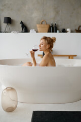 Sensual relaxed woman drinking red wine while taking bath with foam