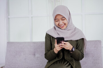 Fototapeta premium A beautiful Asian woman in hijab smiles cheerfully sitting on a sofa using a mobile phone, receiving good news