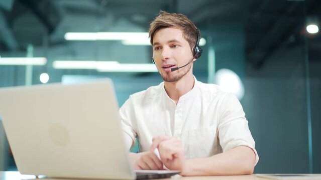 Smiling man call centre operator, customer support agent wears headset consults client online typing on laptop, talk in internet computer chat, helpline operator secretary make conference video call