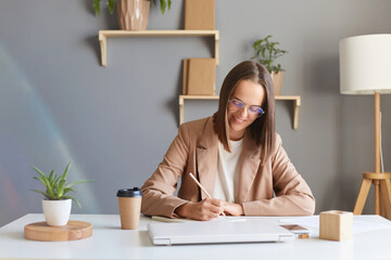 Indoor shot of smiling joyful woman freelancer or designer with brown hair wearing beige jacket working in office, drawing sketches or checking paper documents.