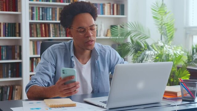 Young focused African American entrepreneur uses laptop and mobile phone to fill out complex financial statements for tax purposes or type important email messages sits at desk in office with bookcase