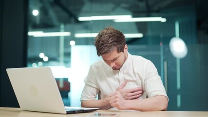 A young employee suffers a heart attack at the workplace. Feeling pain, holding his chest while sitting in modern office at work. IT programmer having cardiac medical health problem. Sick businessman
