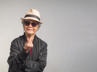 Senior woman in a black jacket wearing sunglasses wearing a hat and hand showing mini heart symbol with a smile while standing on a gray background