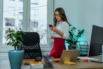 Young woman is checking her schedule on her phone as she drinks coffee in her office