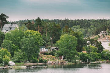 Fototapeta na wymiar Sweden. Beautiful Swedish Wooden Log Cabins Houses On Rocky Island Coast In Summer Day. Lake Or River Landscape.