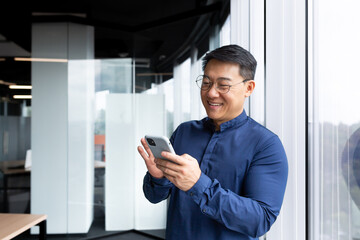 Satisfied with achievement Asian businessman inside office using phone, mature man in shirt near window reading news online from smartphone, successful boss in glasses.