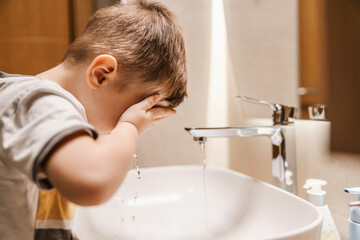 A cute little boy is washing his face in the bathroom.