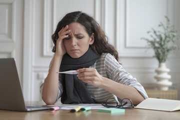 Unhealthy spanish woman wrapped in warm knitted scarf looking anxiously on digital thermometer