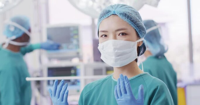 Video portrait of asian female surgeon in cap, gloves and face mask in operating theatre, copy space