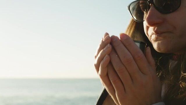A cool Morning at the Sea, a girl warms her hands holding a hot mug with a drink, steam is coming. Close-up.
