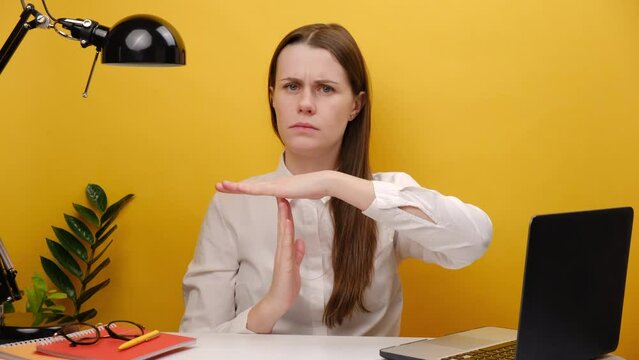 Strict employee young business woman 20s in white shirt sitting work at office desk with laptop holding stop gesture hands perpendicularly, posing isolated over yellow color background wall in studio