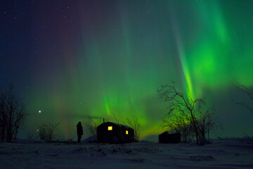 Winter Arctic landscape with the aurora borealis over the snowy tundra. A man stands near the hunting lodge and admires the northern lights. The planets Venus and Jupiter are visible above the horizon