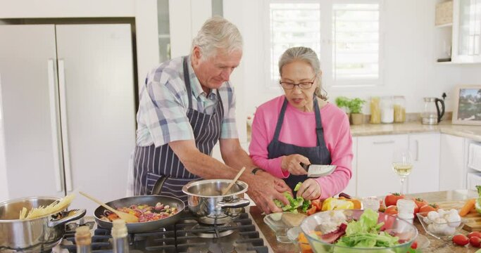 Happy diverse senior couple wearing aprons and cooking in kitchen