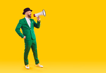 Full body young man in hat and green suit standing on empty blank yellow copy space studio background speaks through megaphone, makes announcement, advertises holiday sale or Saint Patrick's Day party