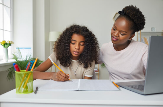 Family doing homework together. Young parent helping child with school assignment. Supportive African American mother and daughter writing and drawing with pencil in notebook at desk with laptop PC