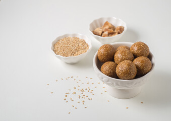 Bowl of sesame seed laddus with raw ingredients on white background. Til laddoo or sesame candy is a traditional Indian sweet balls made with sesame seeds and jaggery mainly during winter season.