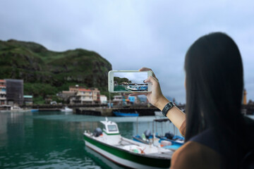 Back view portrait of a woman taking photo of fishing village full of anchored fishing boat in cloudy day by her smart phone 