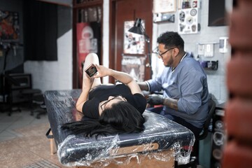A woman is texting with her phone on a stretcher while being tattooed on her leg