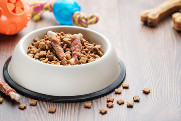 A bowl with dog food, dog treats and toys on a wooden floor.