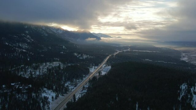 Splendid Aerial Approach Shot of Cariboo Highway 95 Running into the Horizon Surrounded by Grand Mountains Range Covered in Clouds During Warm Sunrise Light: Panoramic View of Winter Landscape