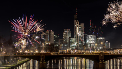 Fototapeta na wymiar New years eve with fireworks above the skyline of Frankfurt - Main at night at a cold day in winter with colorful reflections in the water.