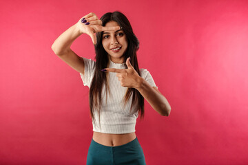 Portrait of young brunette woman wearing white ribbed crop isolated over red background perfectly playful, doing frame gesture with hands and smiling. Frame, recording, archive, camera, movie concept.